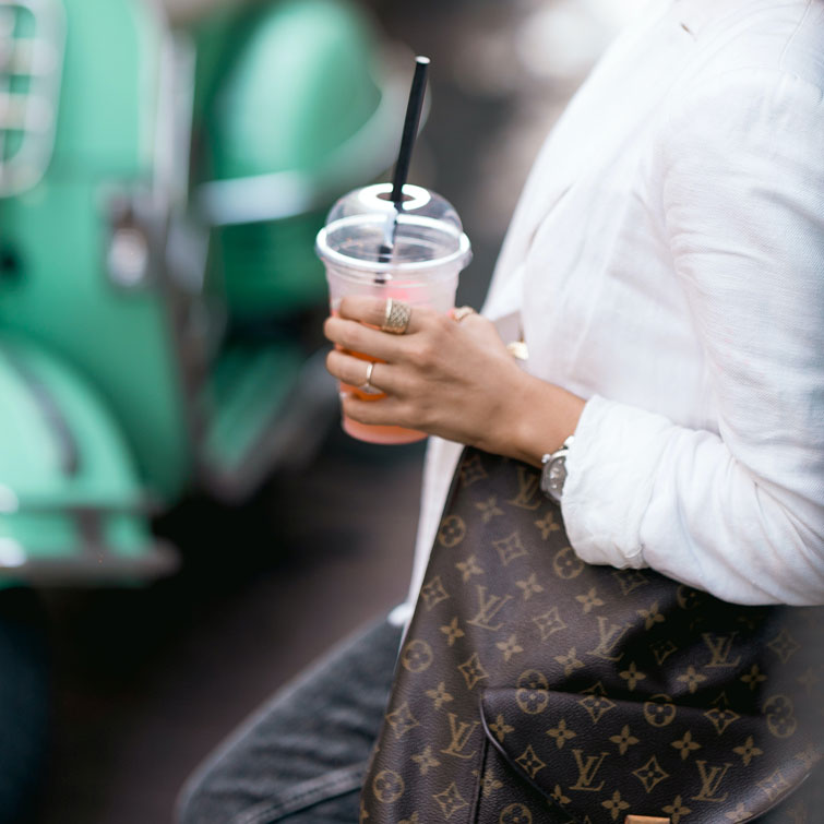 femme qui tient une boisson et qui porte un sac Louis Vuitton marron. Image qui permet d’illustrer comment reconnaître un vrai sac Louis Vuitton.