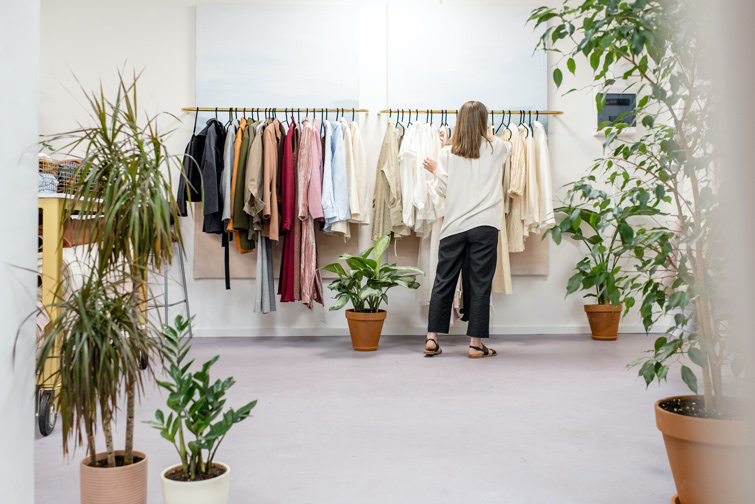 Boutique de mode responsable femme d’un style épuré avec des plantes. Une femme se tient devant les cintres pour ranger des vêtements.